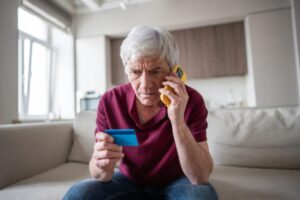uomo anziano al cellulare con il bancomat in mano e l'espressione disperata