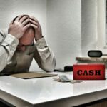 uomo con le mani sulla testa e scritta "cash" sulla scrivania